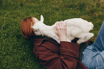 Fototapeta premium Bright outdoor scene shows a human lying on grass with a goat kid resting on chest, displaying affection and bond, a candid meadow moment celebrating simple lifestyle and animal companionship