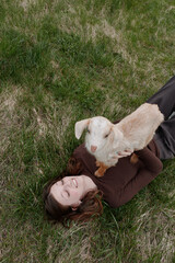Fototapeta premium Relaxed woman lying on grass with a playful goat kid perched on her chest, outdoor lifestyle moment highlighting affection and cuddle, nature and companionship in a gentle, joyful scene.