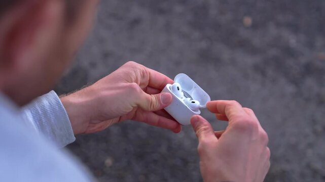 Over shoulder close-up of a young man opening a wireless earbud charging case and removing an earphone in slow motion.