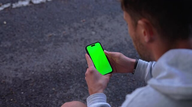 Over shoulder view shows a young man tapping and swiping a green screen smartphone while seated outdoors in slow motion.