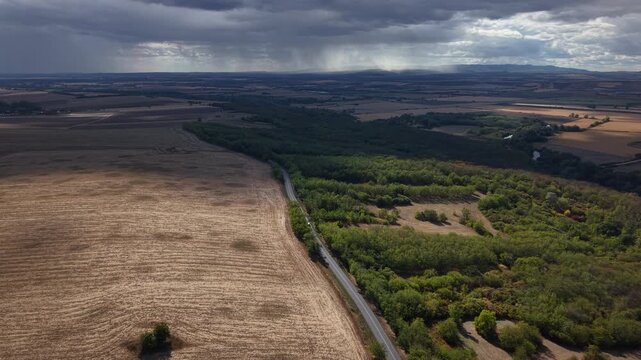 An aerial view of a country road splitting agricultural fields and lush forest under a dramatic, rain-filled stormy sky.