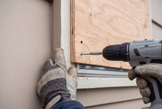Gloved worker uses a cordless drill to secure plywood over a house window, creating temporary protection before severe weather or flooding.