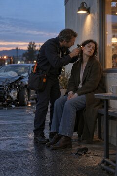 Paramedic examining injured woman with otoscope after car crash on evening street. Wrecked damaged car and emergency responders in background. Roadside medical assistance and emergency care.