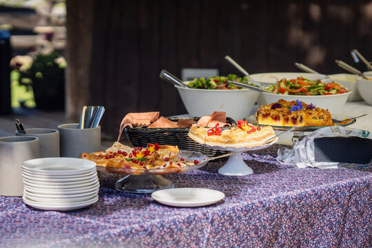 A buffet table displaying an assortment of homemade fruit cakes and pastries decorated with fresh berries and edible flowers, alongside bowls of fresh salads and neatly stacked white plates. 