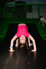 Girl doing a gymnastic bridge pose on a black trampoline with green safety padding indoors.