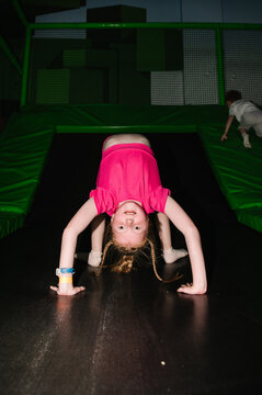 Girl doing a gymnastic bridge pose on a black trampoline with green safety padding indoors.