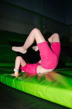 Playful girl performing a back bend stretch on a green trampoline pad in an indoor play center.