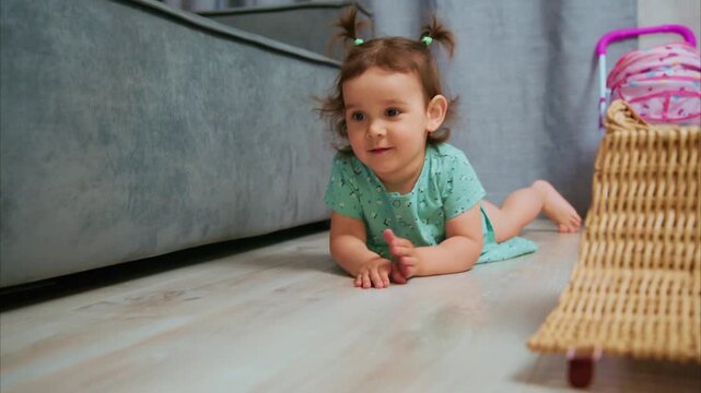 Tiny girl smiles, crawling on hardwood floor. She wears green dress, hair in twin pigtails. Wicker basket sits nearby, with pink toy inside. Child looks toward camera, reaching with open hand