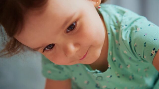Cute baby girl smiles softly, eyes bright and curious. Her curls frame a gentle, sweet expression. She wears a mint green, patterned top. Soft background highlights her innocent, joyful gaze