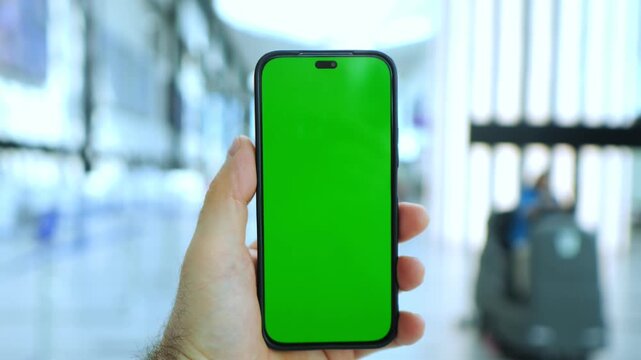 Hand holds smartphone with green screen. Background shows blurred airport terminal. Three keyframes show same scene. No animals or plants present. Focus remains on phone and hand