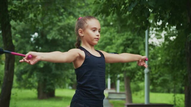 Young girl practices with batons outdoors. Her focused expression shows dedication and concentration. Green trees frame her in a peaceful park setting