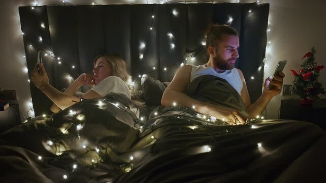 Soft lights drape the bed like stars. Couple sits side by side, absorbed in phones. Night air feels warm and quiet. Dark headboard glows with twinkling lights. Their faces show calm