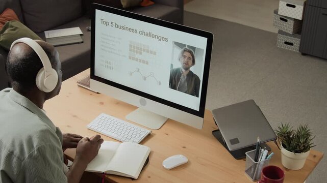 Mature African American man wearing headphones taking notes while watching business webinar on computer. Perfect for corporate training, e-learning platforms, remote work promotions