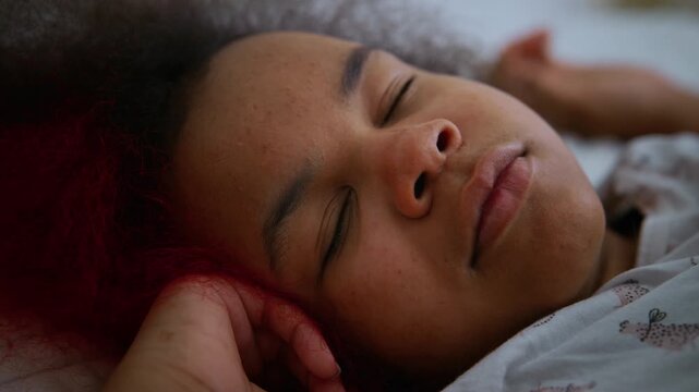 Softly sleeping child rests peacefully. Curls frame a serene, dreamy expression. Hand gently cradles ear, eyes closed. Bedding soft and warm beneath. Tranquil moment of quiet rest