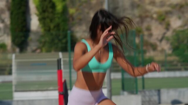 Female athlete adjusting posture while walking on outdoor sports court in sunny weather, wearing turquoise sports bra and pastel shorts, hands moving from forehead toward hips