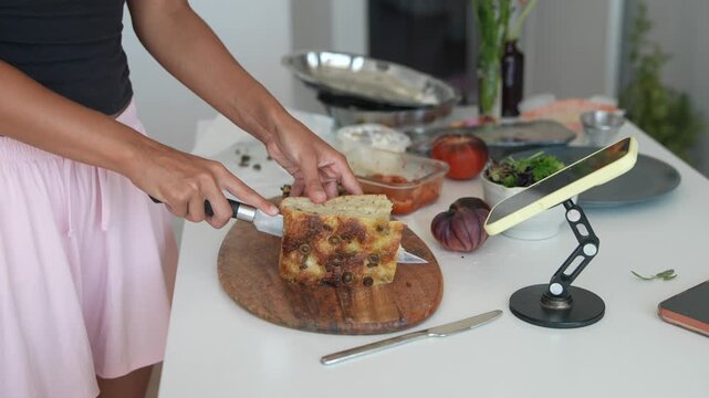 Hands slicing olive focaccia on round wooden cutting board in home kitchen with chef knife at countertop level under soft daylight