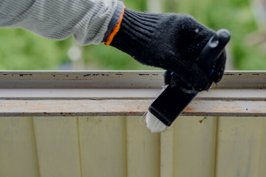 Closeup of gloved hand using small brush to remove dust from metal balcony frame before painting, highlighting careful cleaning motion, textured surface, outdoor setting and protective glove