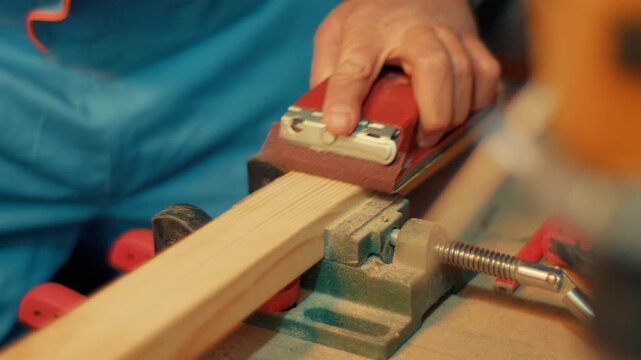 Slow motion close-up of hands using a red sanding block to smooth a wooden plank clamped in a vise. Authentic DIY woodworking footage for craft, carpentry and maker projects