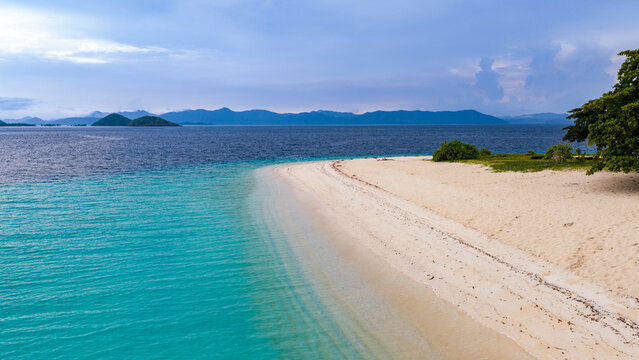 Aerial view of a pristine white sand beach meeting turquoise waters, with lush green trees on the shore and distant mountains under a cloudy sky in Busuanga, Mimaropa, Philippines.