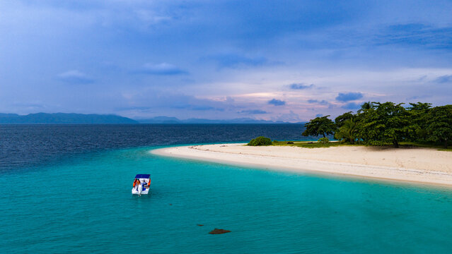 Aerial view of a small boat floating in turquoise water near a white sand beach with lush green trees and distant hills under a cloudy sky in Busuanga, Mimaropa, Philippines.