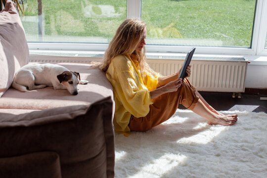 Woman sitting on living room floor reading a tablet beside a resting dog. Relaxed home leisure and digital reading. Pet companionship and everyday domestic life. Cozy indoor lifestyle