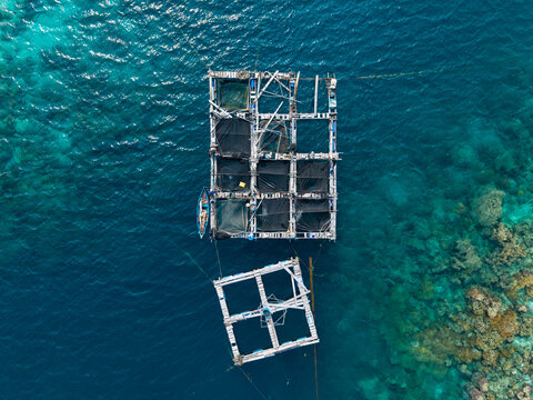Aerial view of fish cages floating in clear turquoise water with a small boat nearby and coral reefs visible below in Karimunjawa, Central Java, Indonesia.