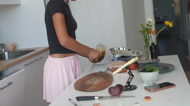 Young woman arranging ingredients near foil tray and salad bowl on bright kitchen countertop, hands reaching in modern home cooking setup with cutting board, knife, and packaged bread in foreground