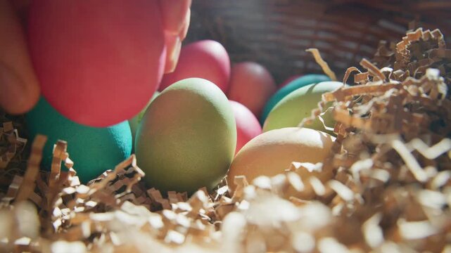 Hand gently picks up a red egg from a basket. Colorful eggs rest among shredded paper and soft light. Warm tones suggest springtime joy and celebration. Perfect for Easter, parenting. in this scene