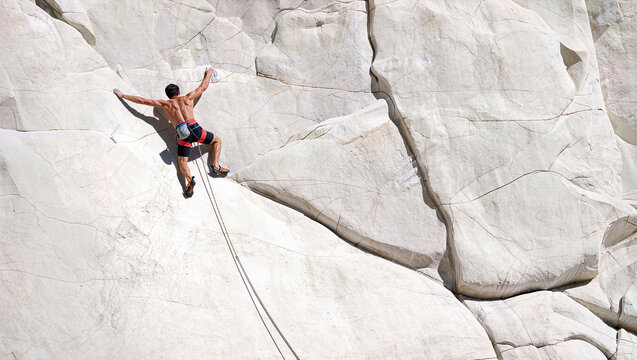 Lone climber scaling vast white limestone cliff with trailing rope under hard sunlight, wide view of man on smooth cracked rock face, conceptual solo challenge and fearless determination concept