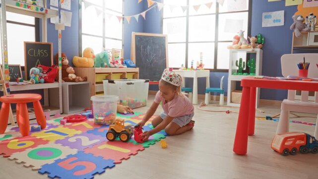 Little girl wearing tiara plays with toy truck on foam number mat in daycare building; childhood joy.