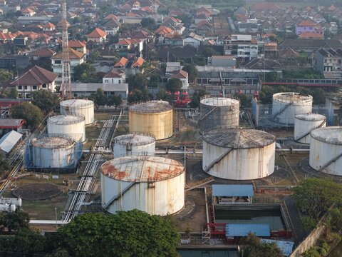 Aerial view of PT Pertamina Terminal BBM Sanggaran with industrial fuel storage tanks, pipelines, and residential rooftops in Kecamatan Denpasar Selatan, Bali, Indonesia.