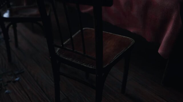 Empty wooden chairs sit near a table covered by a pink cloth inside a room with low light during the evening hours. The floor is made of dark wood planks and the atmosphere remains quiet and still.