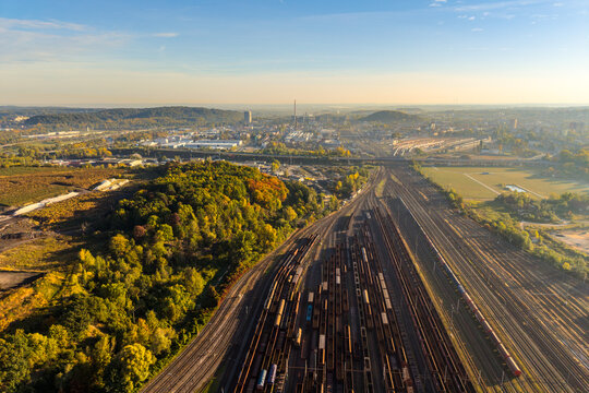Aerial view of a large wagon depot with numerous freight trains on parallel tracks, surrounded by autumn forests and an industrial cityscape in Czechia.