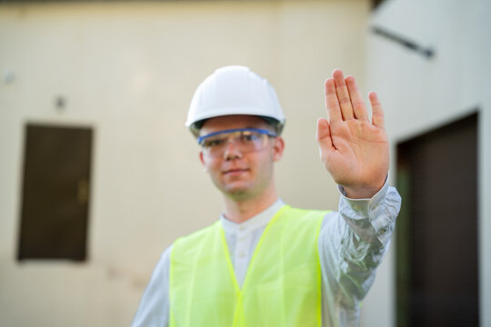 Construction worker in hard hat and high visibility vest raising hand with stop gesture outdoors. Workplace safety communication and hazard prevention