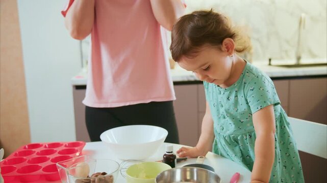 Little girl mixes batter with spoon in kitchen. Mother guides her gently with bowl and whisk. Child focuses on cooking with curious expression. Warm home scene, perfect for parenting or family content