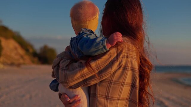 A woman holds her bundled baby on a beach at golden hour beside the calm ocean