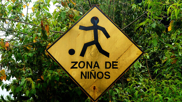 Letrero de zona de ni&ntilde;os en espa&ntilde;ol junto a cerco de madera y follaje verde, se&ntilde;al de advertencia con figura y bal&oacute;n, prevenci&oacute;n de accidentes, conducci&oacute;n responsable y cuidado de la infancia