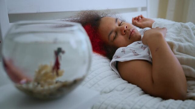 Young woman sleeps peacefully beside fishbowl. Red hair rests gently on white bedspread. Fish swims calmly in clear, round container. Soft lighting enhances tranquil bedroom atmosphere