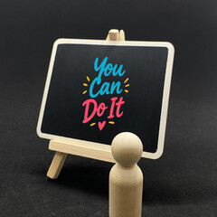 Inspirational mental health photo featuring a wooden peg figure facing a miniature blackboard on an easel with colourful handwritten quote You Can Do It on a dark background. © izzy sabki
