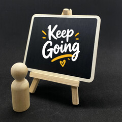 Inspirational mental health photo featuring a wooden peg figure looking at a miniature blackboard on a easel with handwritten quote Keep Going on a dark background. © izzy sabki