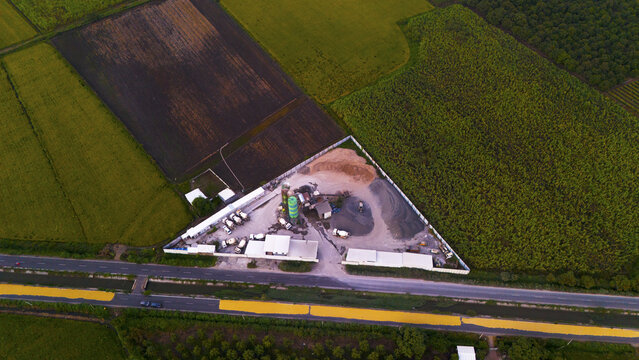 Aerial view of a concrete batching plant with silos and trucks situated in a triangular plot surrounded by agricultural fields and a road in Surat, Gujarat, India.