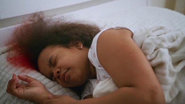 Girl sleeps peacefully in bed, wrapped in white sheets. Red highlights frame her curly hair, soft and natural. Her eyes are closed, face relaxed, body at ease. Ideal for scenes of rest, comfort