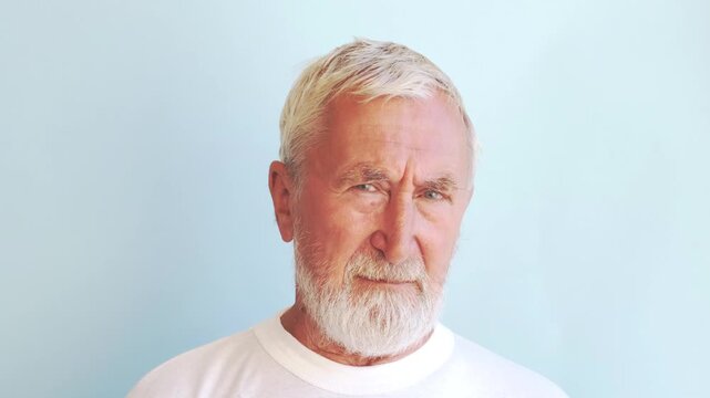 Skeptical senior man raising eyebrow, grey bearded elderly man in white t-shirt looking at camera with doubt on light blue background. Distrust and doubt concept, studio close up