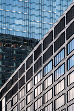 Diagonal perspective of a light colored office building with repeated square windows against a taller glass tower creating contrasting layers of modern urban architecture