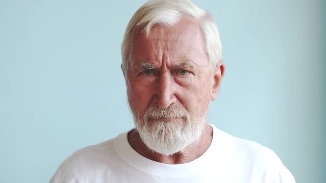 Angry senior man frowning at camera, grey bearded elderly man in white t-shirt with displeased face on light blue background. Displeasure and grumpy old man concept, studio close up