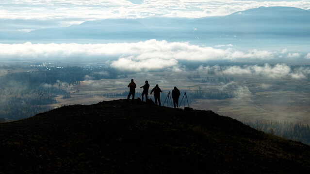 Panoramic aerial view of landscape photographers with tripods capturing majestic misty mountains from a high summit at dawn.