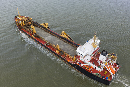 Aerial view of a trailing suction hopper dredger navigating through the sea with a full load of sand in its hopper and industrial deck machinery visible, United Kingdom.