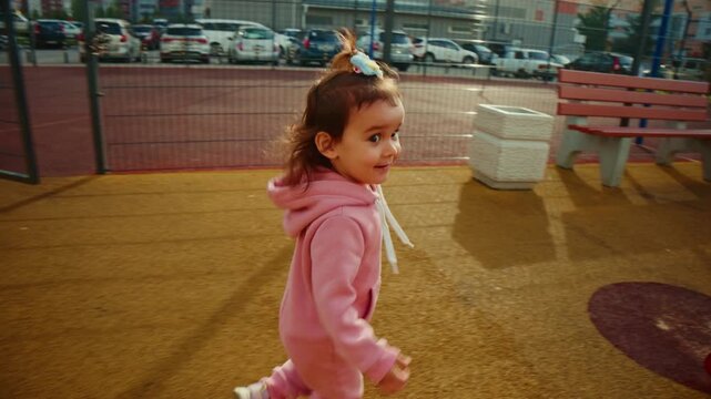 Little girl in pink hoodie walks on playground surface. Curly hair bounces as she moves toward colorful play equipment. Soft sunlight glows on her focused, curious expression