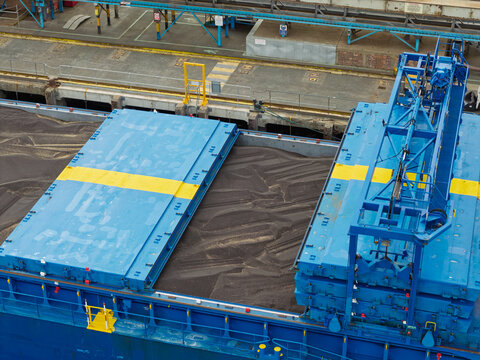 Aerial view of a cargo ship deck with open hatches revealing bulk cargo, featuring blue hatch covers with yellow markings and industrial port infrastructure in the United Kingdom.