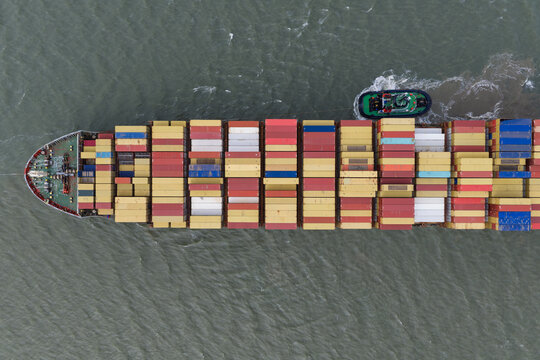 Aerial view of a large container ship loaded with colorful shipping containers being guided by a small tugboat in the United Kingdom.